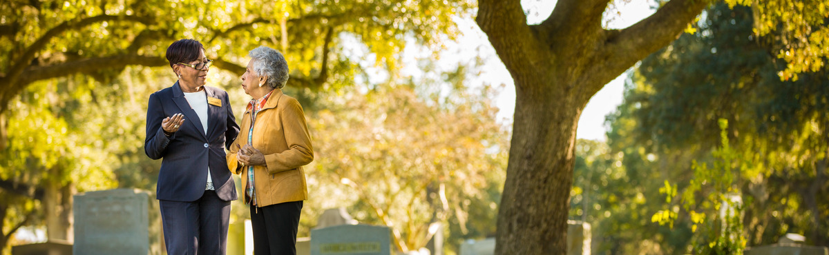 A Dignity Memorial counselor talks with an older woman in a cemetery with a large tree in the background.