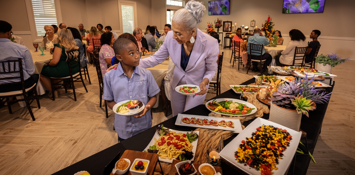 A grandmother and grandson serve themselves at a catering table while attending a funeral reception